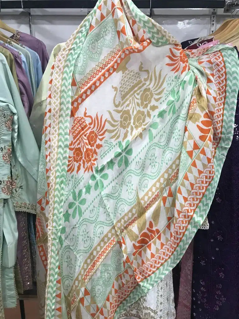 A lightweight printed scarf or shawl draped over a hanger in a clothing store. The fabric has a white base with a multicolor pattern of orange, green, and beige floral and geometric motifs, with a solid orange border.