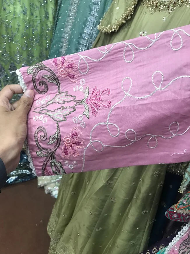 A close-up of a pink embroidered fabric sleeve or garment piece being held by a hand, showing floral and scroll embroidery in pink, silver, and white. Other green and multicolored embroidered garments are visible in the background.