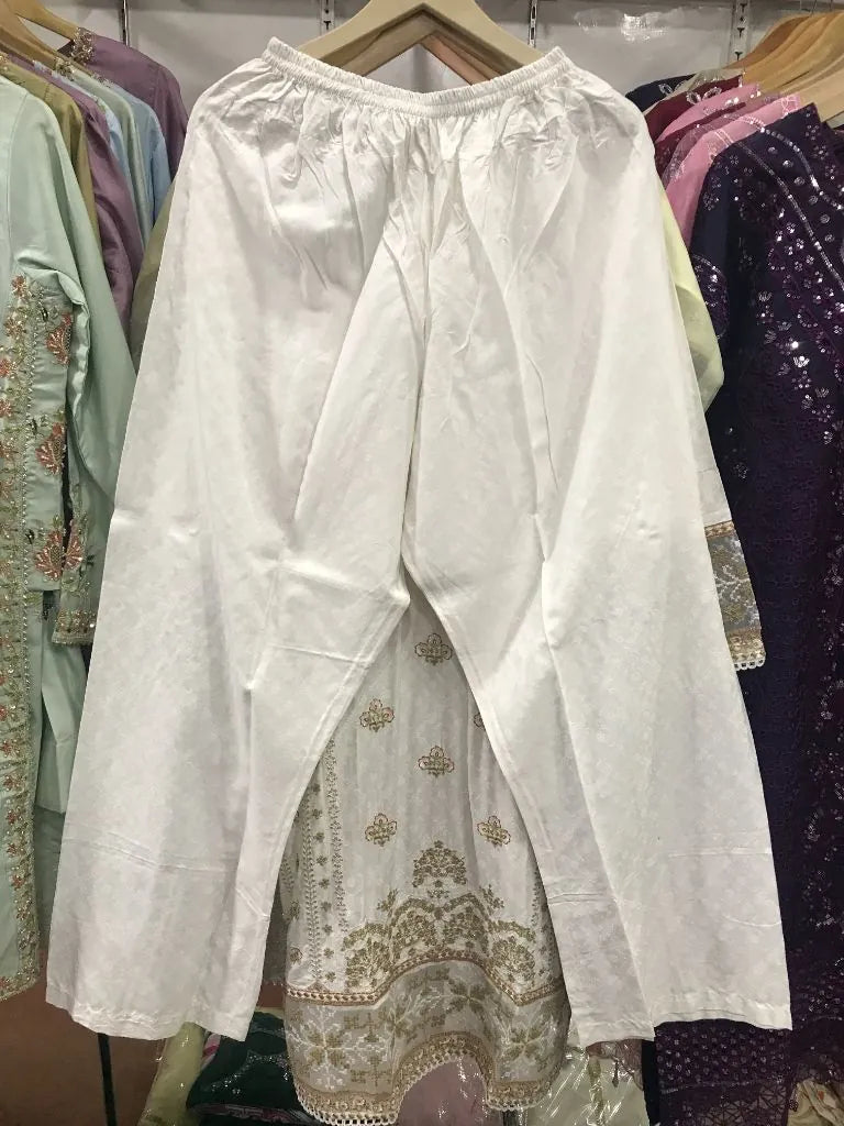 A pair of loose white cotton pants with a drawstring waist, hanging on a wooden hanger in a clothing store. Other colorful garments are visible in the background.