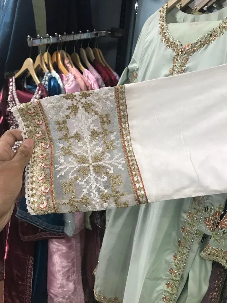 A close-up of a white embroidered sleeve or garment piece being held by a hand, showing detailed gold and beige floral embroidery with a decorative border. Other garments are hanging on racks in the background.