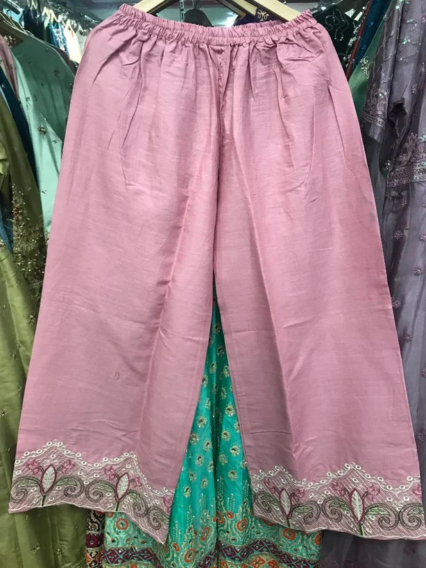 A pair of loose, wide-leg cropped pants in dusty pink fabric with a gathered elastic waistband and decorative embroidered lace trim at the hem, displayed on a hanger in a clothing store.