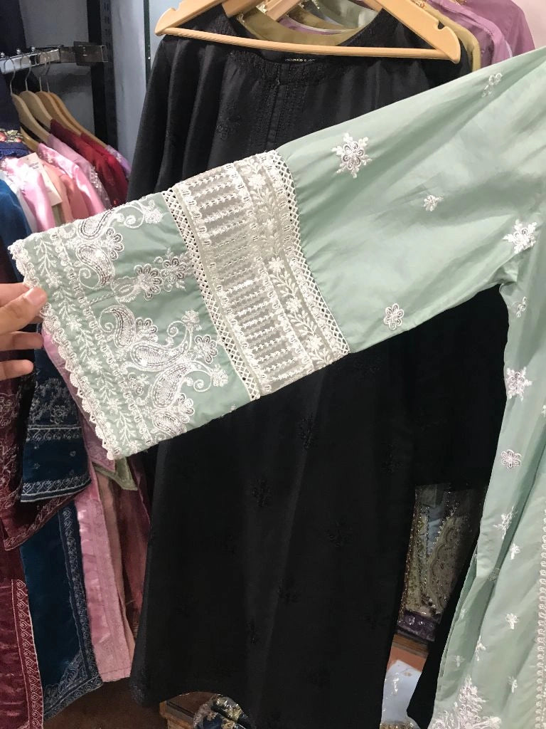 A sage green traditional embroidered garment with white lace and floral embroidery on the sleeve, held up by a hand in front of a black dress on a clothing rack.