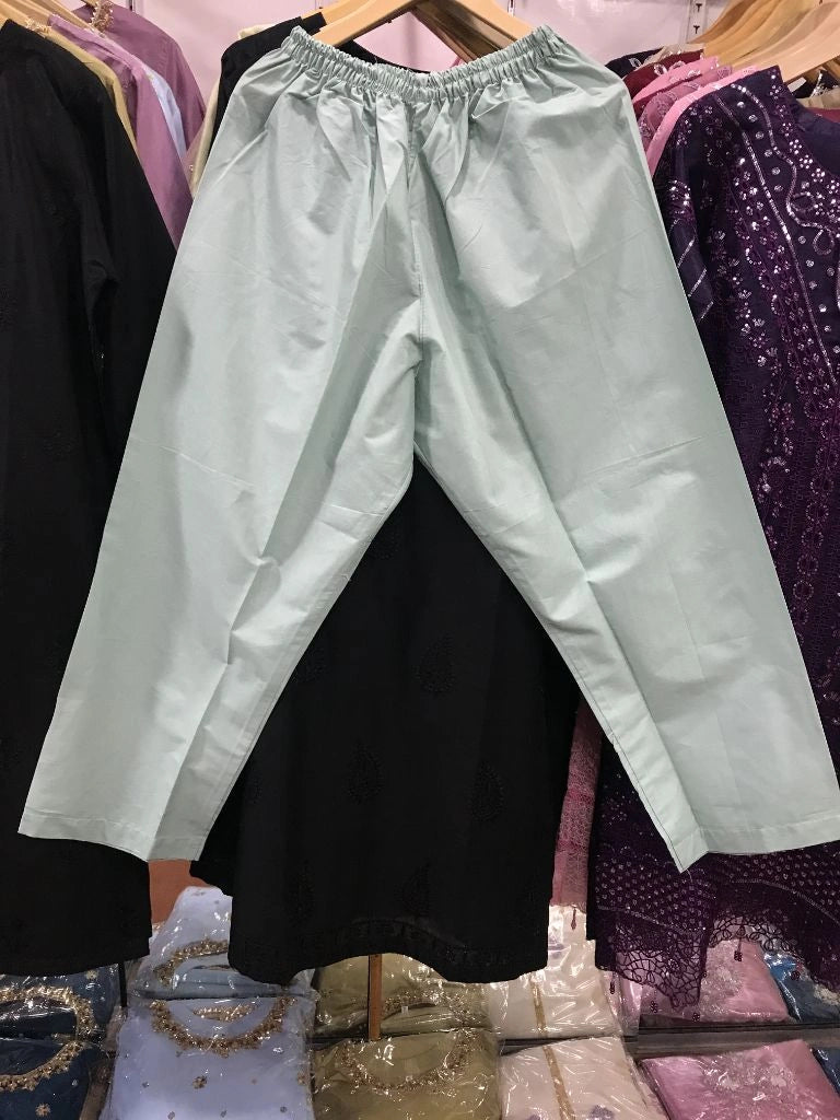 A pair of light sage green wide-leg pants with an elastic waistband, hanging on a wooden hanger in a clothing store. Black and purple garments are visible in the background on hangers.